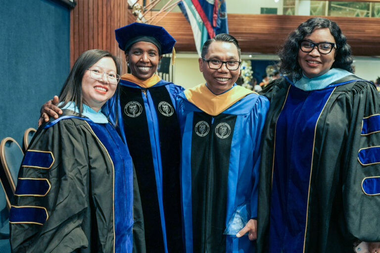 Four graduates in academic regalia celebrating at a ceremony, smiling and posing together.