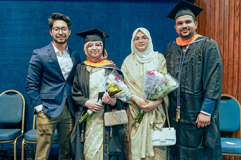 Group celebrating graduation with caps, gowns, and bouquets, standing against a blue background, smiling.