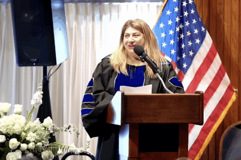 A woman in academic regalia speaks at a podium with an American flag in the background and flowers nearby.