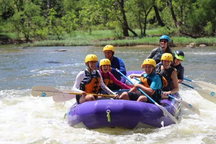 Group of happy people river rafting in a purple raft, wearing helmets and life jackets, enjoying outdoor adventure.