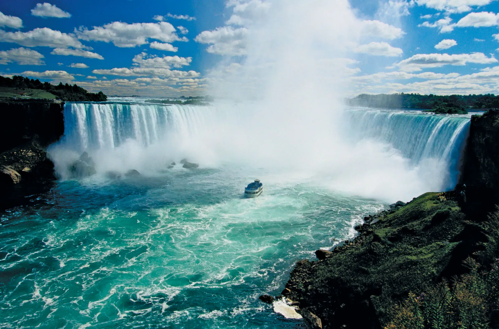 Boat near the misty Niagara Falls under a vibrant blue sky with scattered clouds, capturing the waterfall's power and beauty.