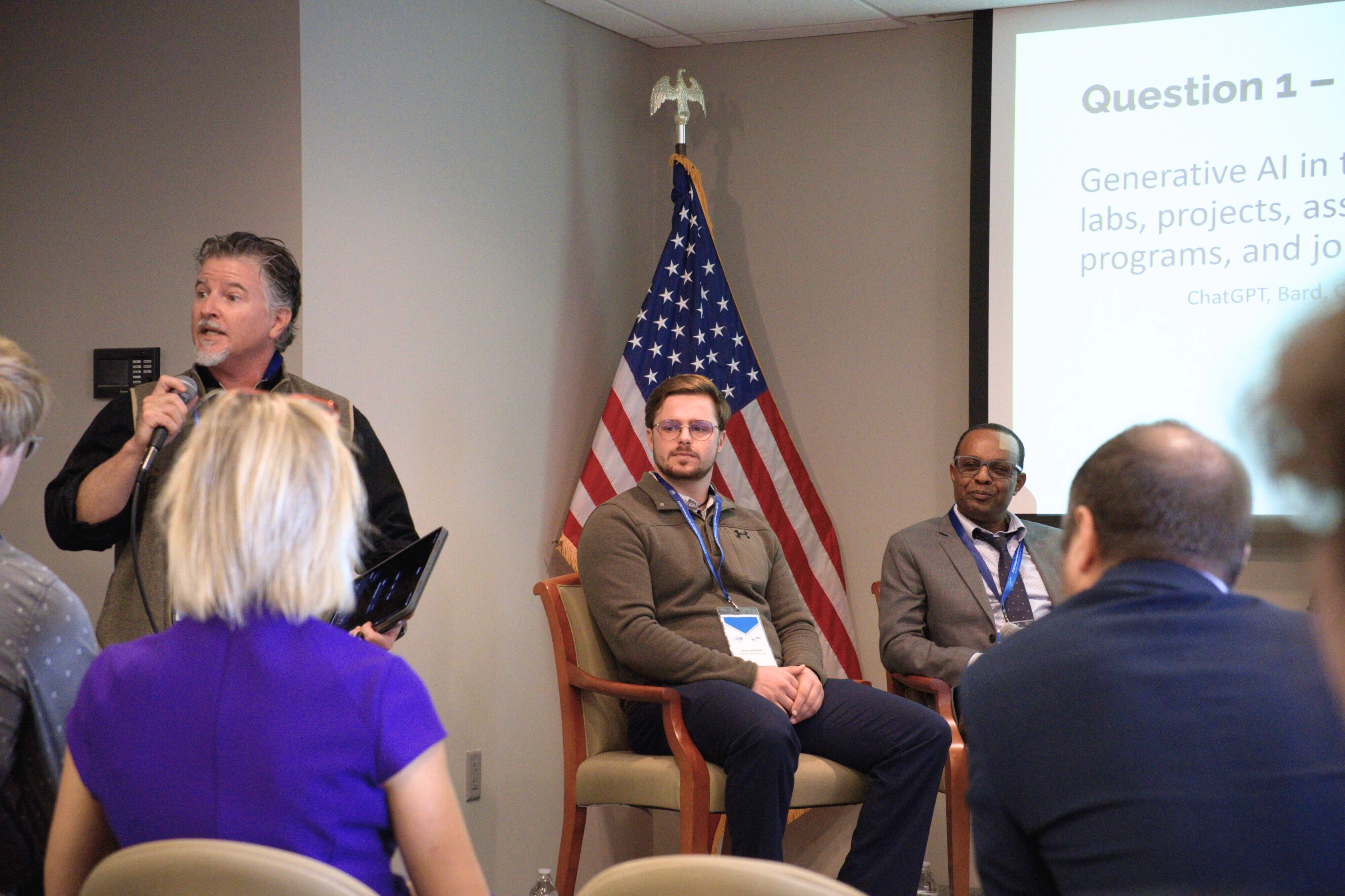 Speaker addressing audience at an AI panel discussion with two panelists, seated near an American flag.