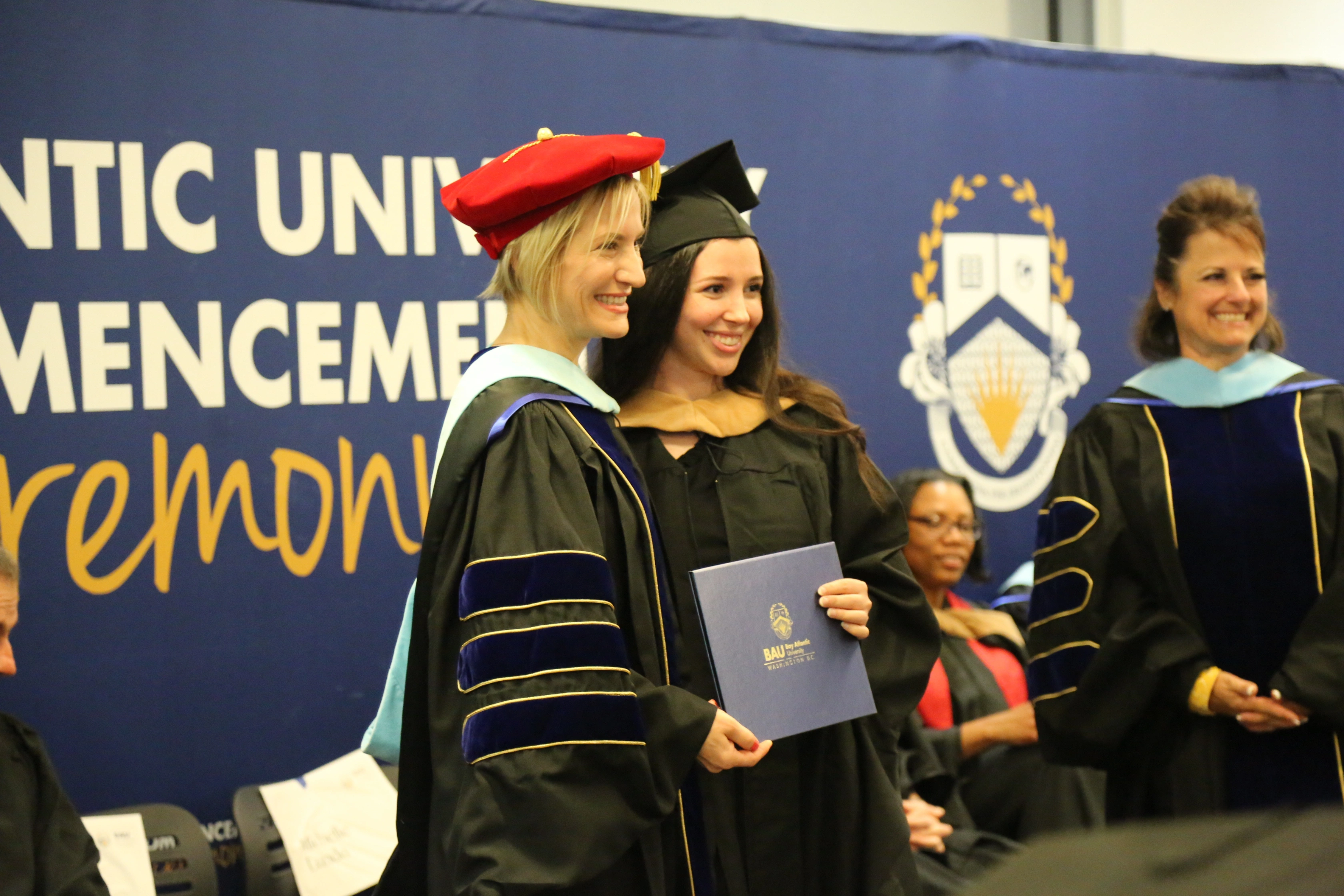 Graduate receiving diploma at university commencement ceremony, smiling with professors, wearing cap and gown.
