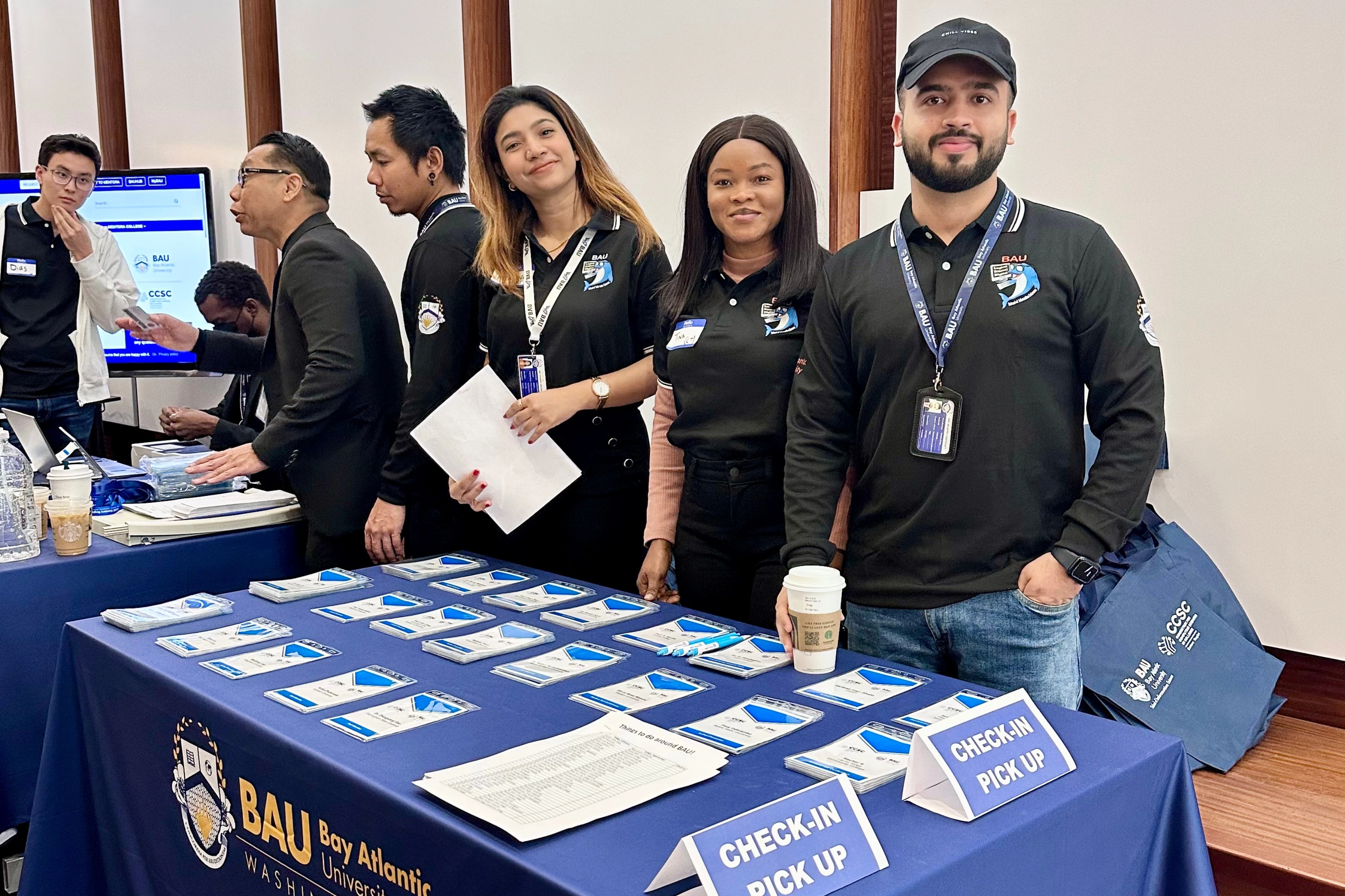 People at a university check-in table with ID badges and signs, volunteering and assisting visitors.