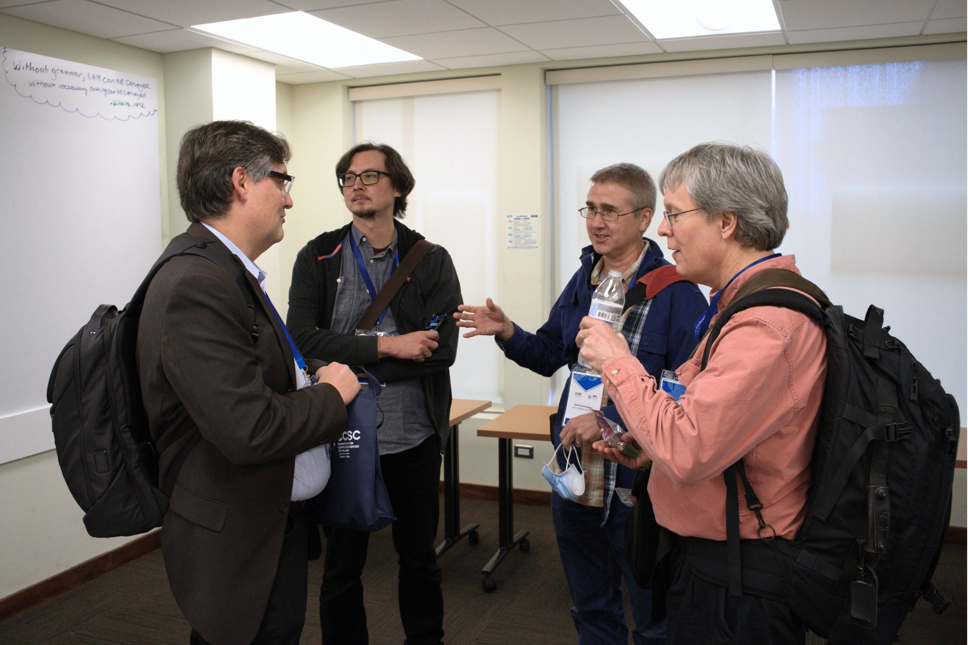 Four people having a discussion at a conference, wearing lanyards and carrying bags in a meeting room.