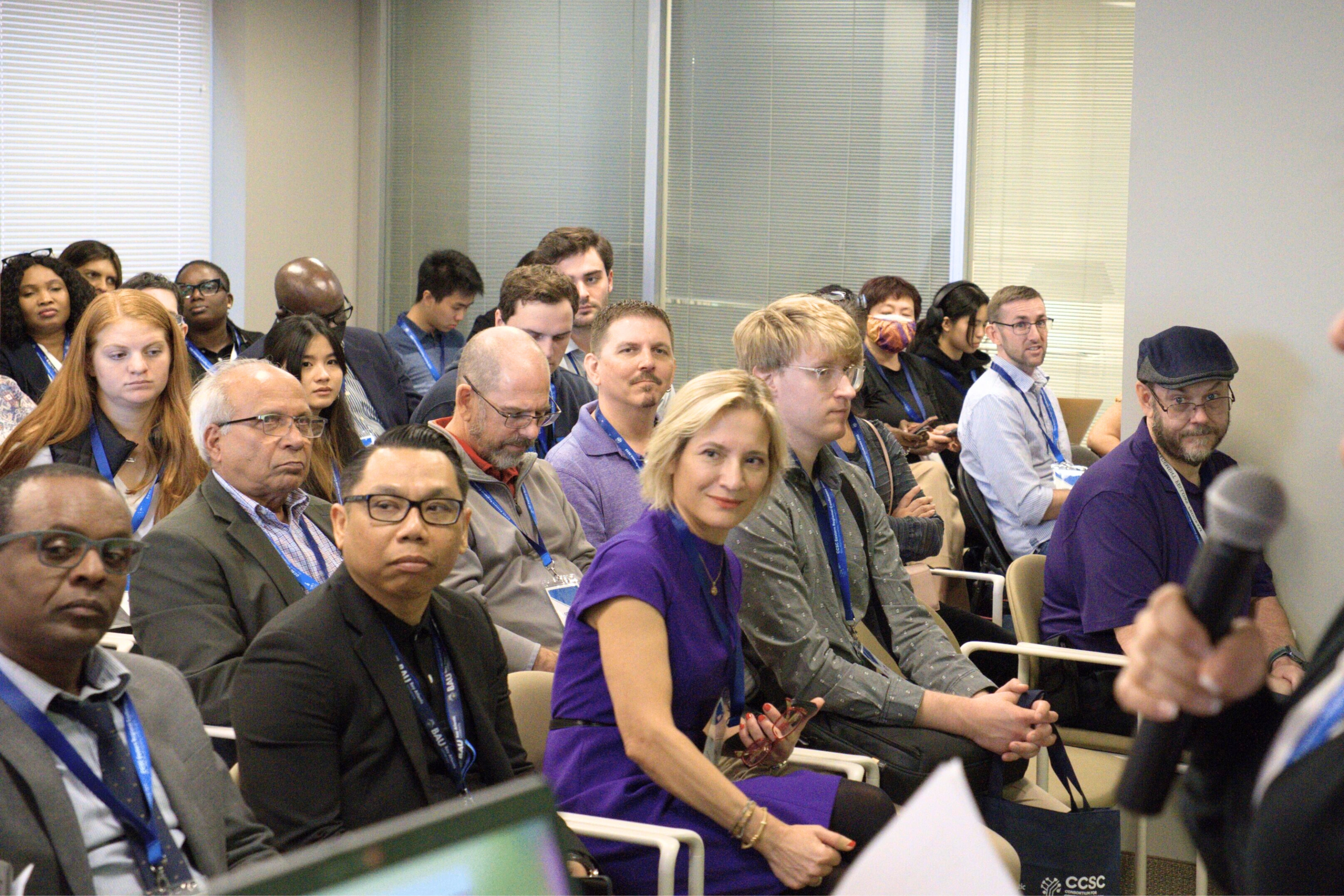 Audience attentively listening to a speaker at a conference, diverse group with lanyards in a meeting room.