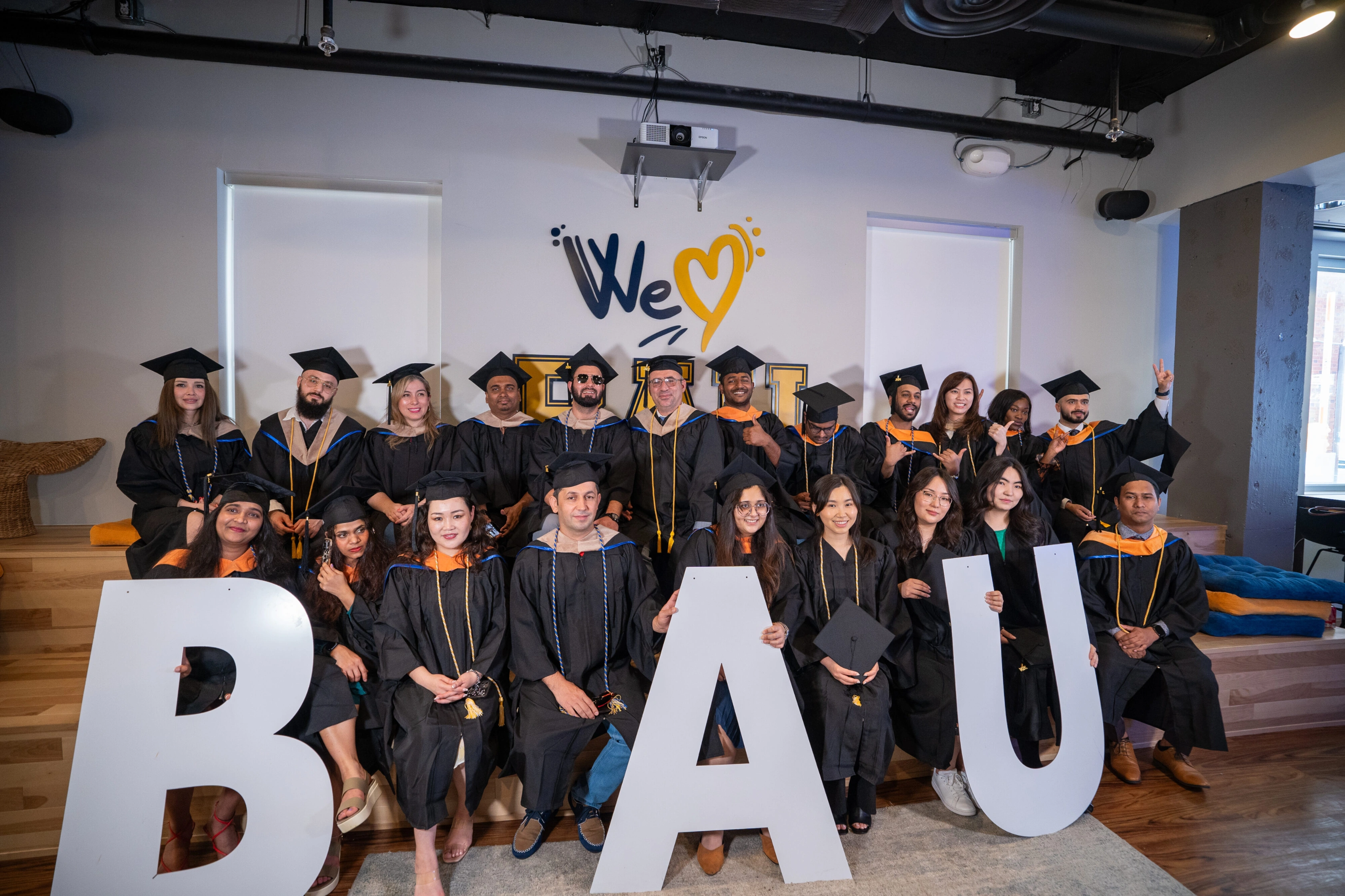 Graduates celebrating at University ceremony, holding large letters, in caps and gowns with We Heart You sign.