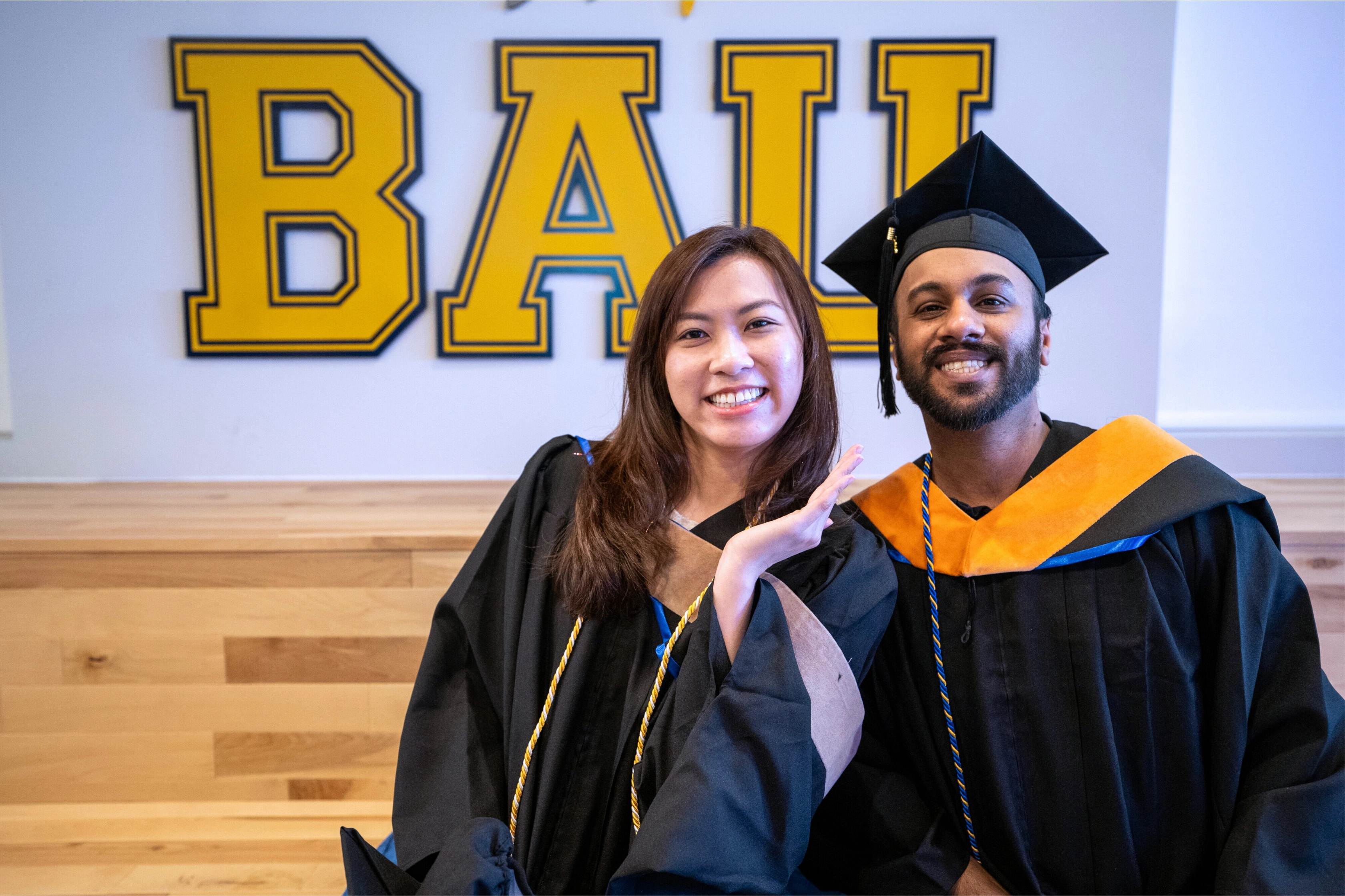 Graduates smiling in caps and gowns in front of yellow BALL sign, celebrating their achievement.
