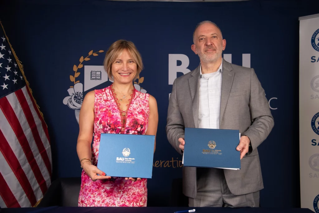 Two people holding certificates at a BAU Washington D.C. event, smiling in front of the American flag.