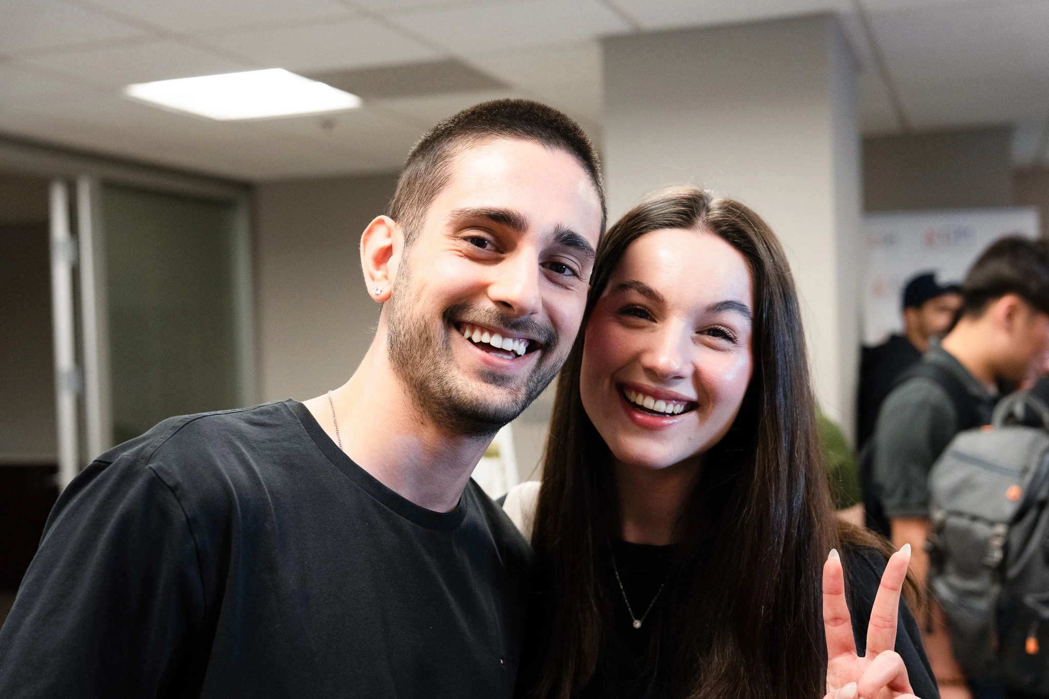 Smiling individuals at an event, one showing a peace sign gesture, enjoying a joyful moment indoors.