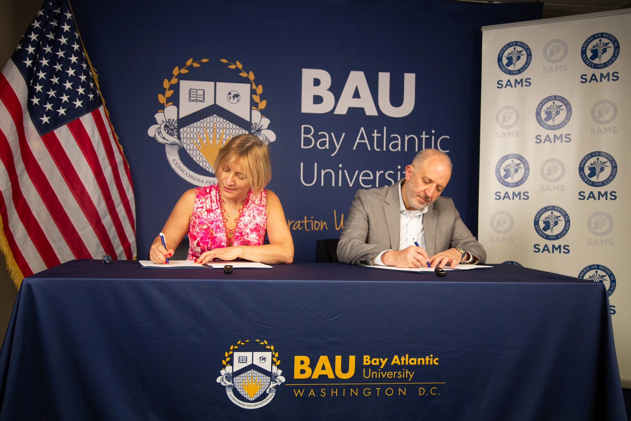 Two people signing a partnership agreement at Bay Atlantic University event with SAMS banner in background.