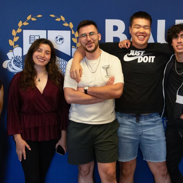 Group of five diverse students smiling in front of a university logo banner, showing camaraderie.