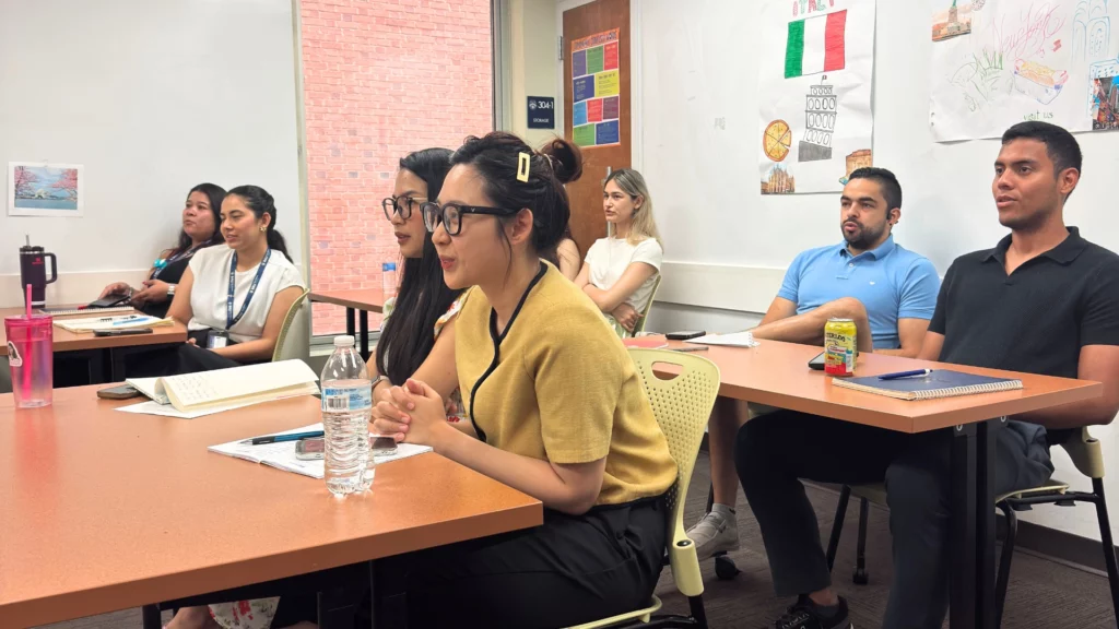 Students attentively listening during a classroom lecture, with notebooks and drinks on their desks.