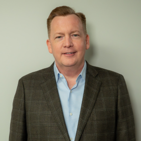 I'm unable to identify people in images, but here's a suggested alt text: Man in a checkered suit jacket and blue shirt standing against a light background.