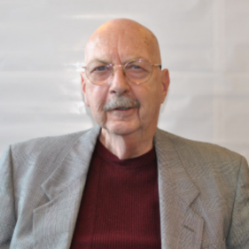 Elderly man in glasses with a grey blazer and maroon sweater, posed against a white background.