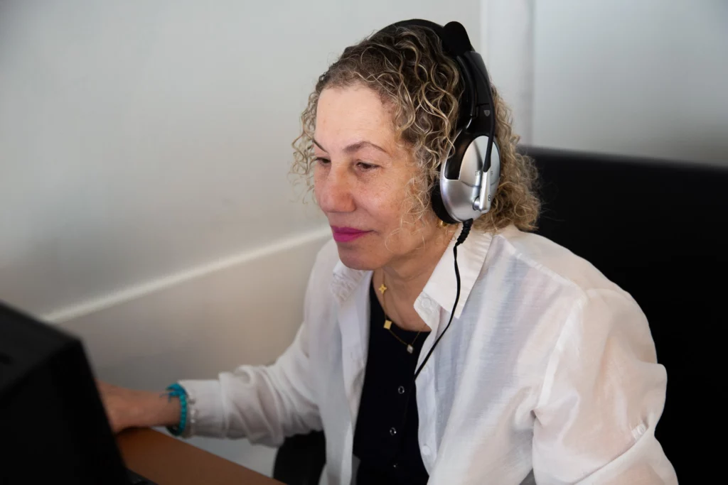 Senior woman with headphones working on a computer, focused expression, white shirt, professional setting.