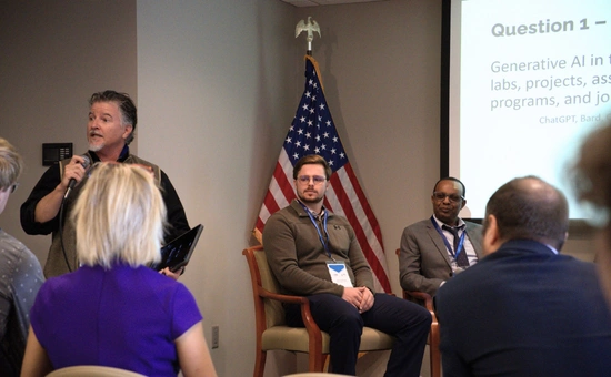 A speaker addresses an audience at an AI conference, with two panelists seated near an American flag and presentation screen.