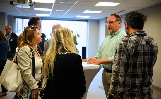 A group of professionals networking at a business event, standing around a table in a bright conference room.