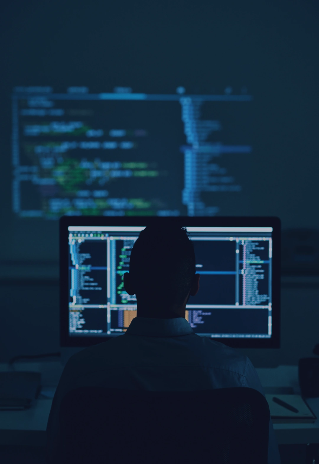 Person coding on dual monitors in a dimly lit room, displaying colorful code and scripts on the screens.