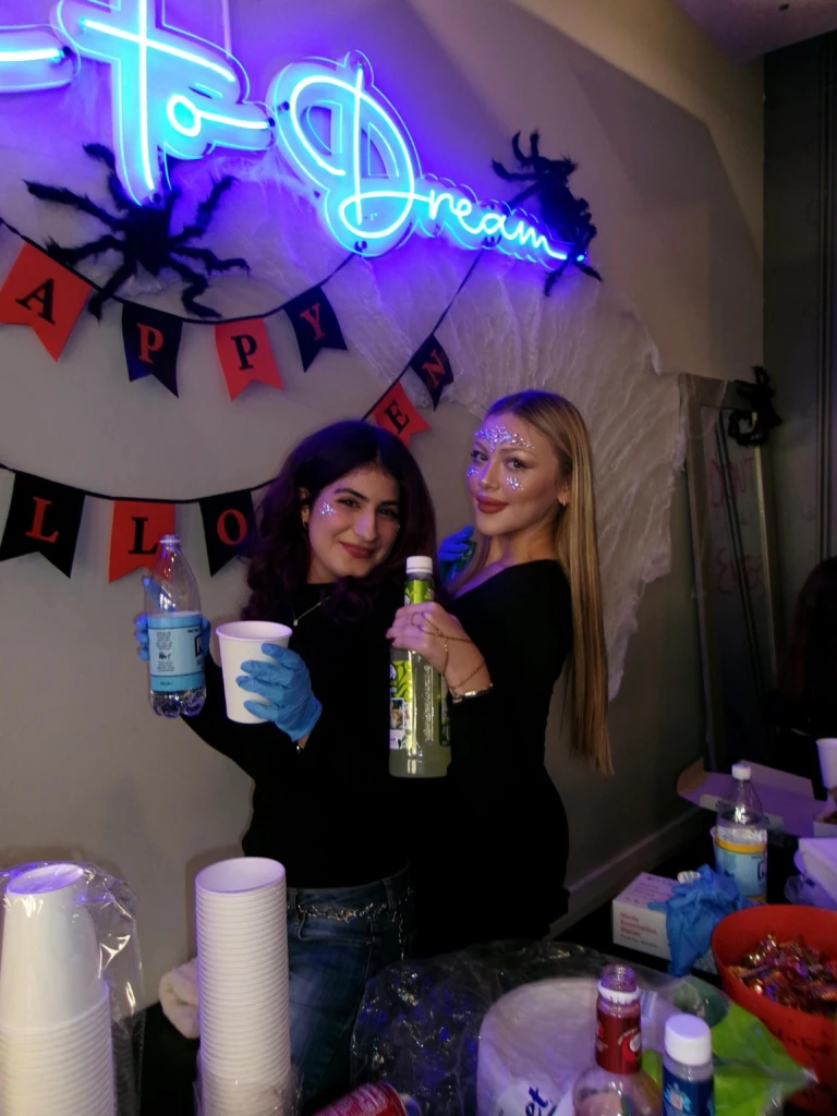 SGA Halloween Party 2025 Two women celebrating at a Halloween-themed event with neon signs, drinks, and decorations in the background.