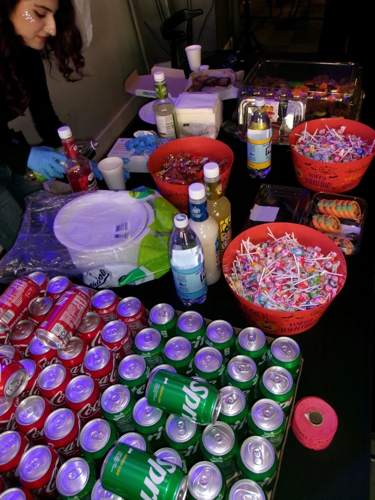 SGA Halloween Party 2025 Party table with assorted sodas, candies, cups, and drinks. Person setting up colorful Halloween-themed refreshments.