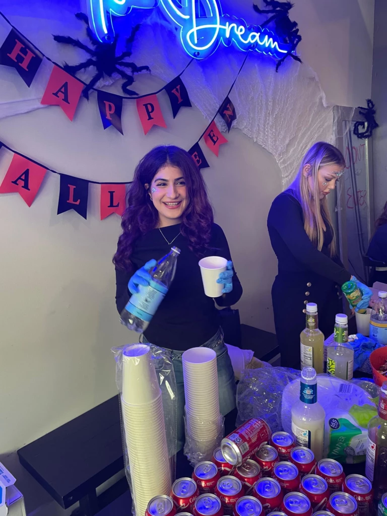 SGA Halloween Party 2025 Woman serves drinks at Halloween party with festive decorations, smiling.
