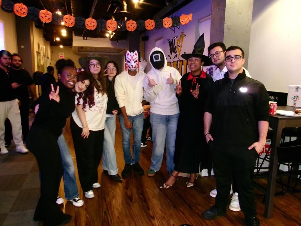 SGA Halloween Party 2025 A group of friends in Halloween costumes posing at a party with festive pumpkin decorations.