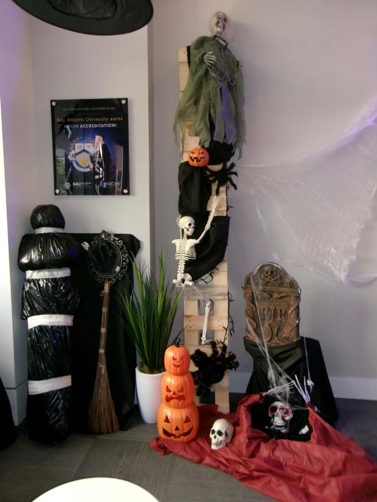 SGA Halloween Party 2025 Halloween-themed office corner with skeletons, pumpkins, a broom, and a tombstone for spooky decoration.