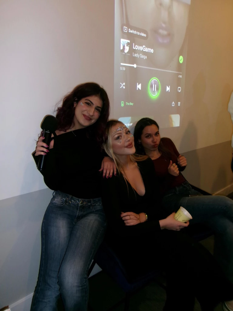 SGA Halloween Party 2025 Three friends at a party, enjoying music with a karaoke mic and a playlist projected on the wall.