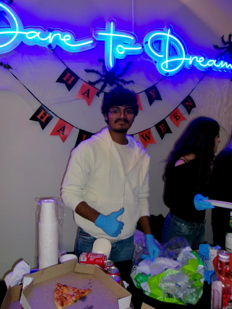 SGA Halloween Party 2025 Man in white hoodie with blue gloves at Halloween party; Dare to Dream neon sign above.