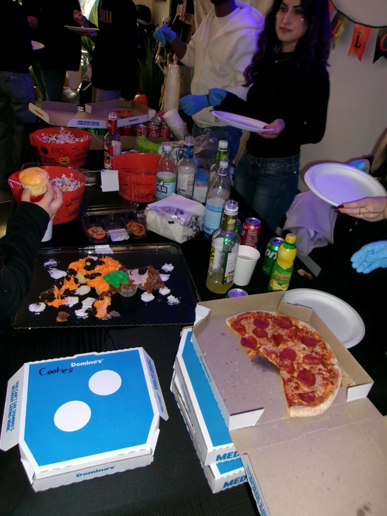SGA Halloween Party 2025 Halloween party with pizza, cookies, drinks, and decorated treats on a table. People serving food and drinks.