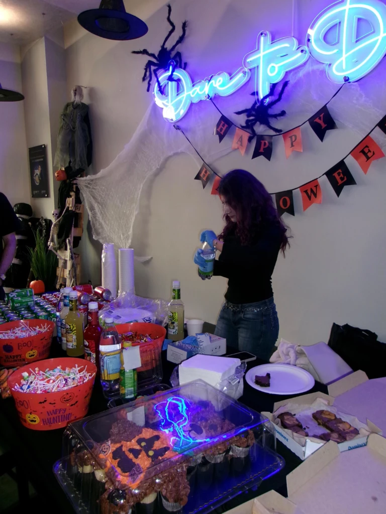 SGA Halloween Party 2025 Halloween party setup with decorations, snacks, and drinks on a table, neon Dare to sign and cobweb decorations.