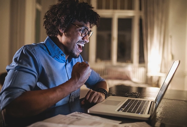 Smiling man celebrates success while looking at laptop, sitting at a desk in a warmly lit room.