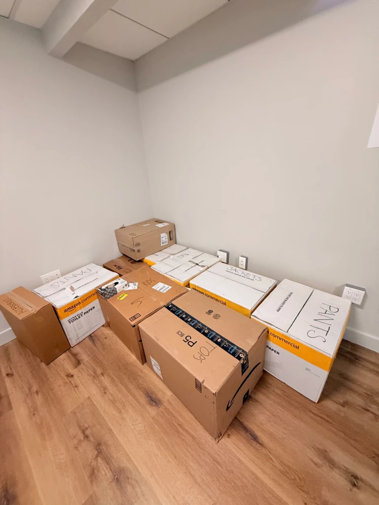 SGA Clothing Donations 2025 Stacked cardboard boxes in a room corner labeled for moving or storage, placed on wooden flooring.