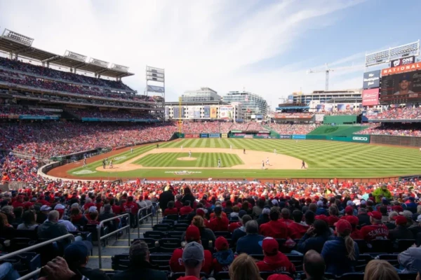 Crowded baseball game at Nationals Park, sunny day, fans in red attire, players on the field, cityscape in background.