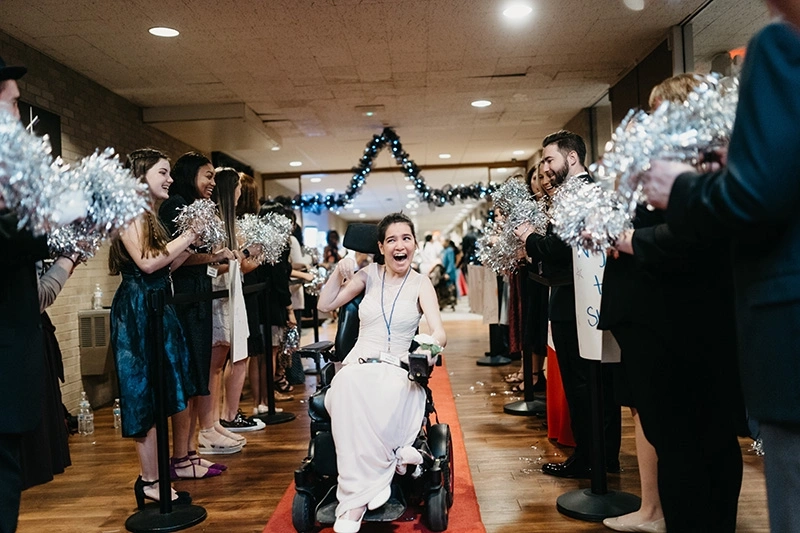 Person in a wheelchair on a red carpet, greeted by cheering crowd with pom-poms at a festive event.