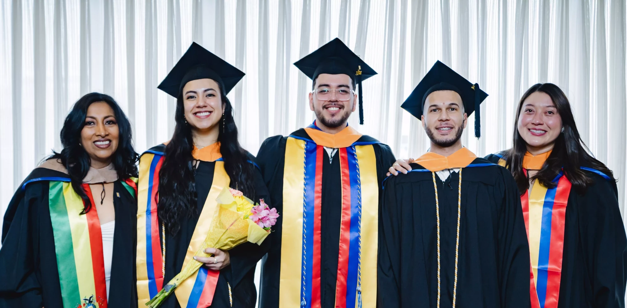 Group of graduates in caps and gowns celebrating with smiles and flowers.