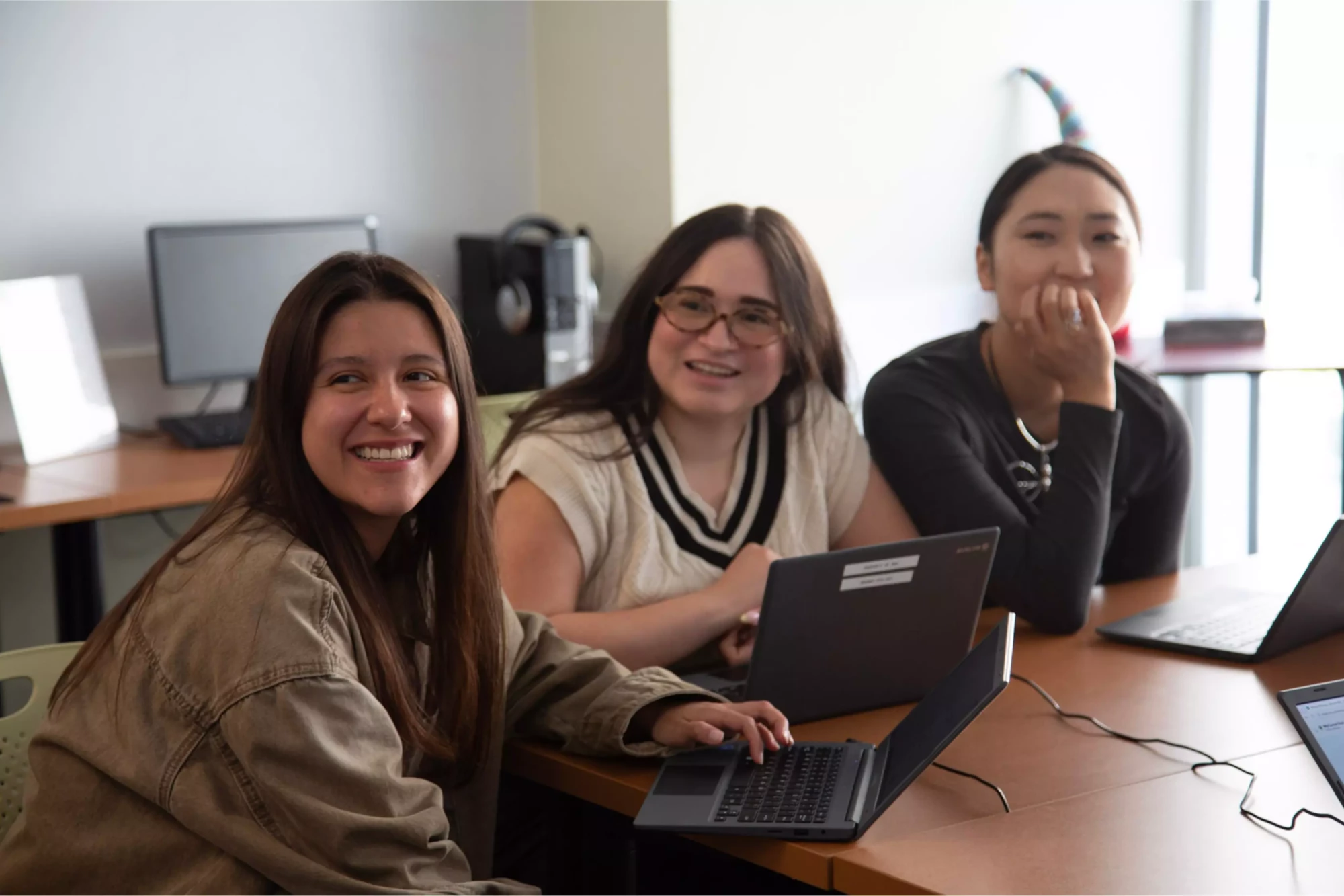 Three women engaging with laptops in a bright office, smiling and discussing ideas.
