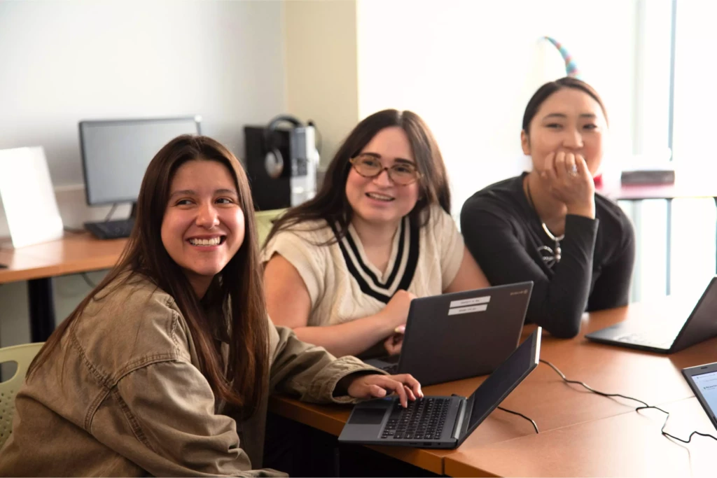 Three smiling women collaborate using laptops in a classroom setting, fostering teamwork and learning.