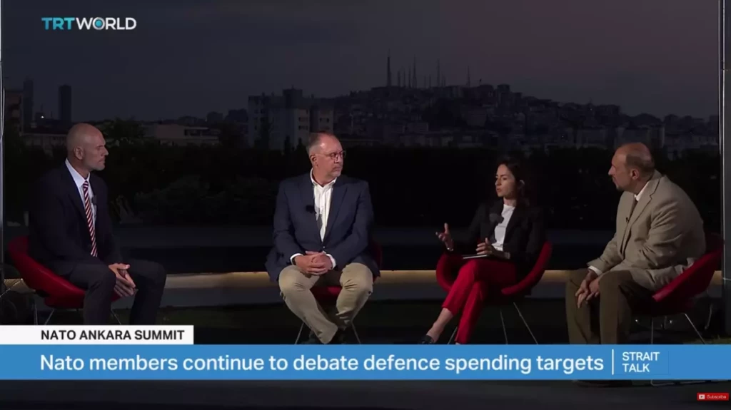 A wide shot of a four-person panel discussion on TRT World's Strait Talk broadcast during the NATO Ankara Summit. The group is seated outdoors at night with a city skyline in the background, and the on-screen graphic reads "Nato members continue to debate defence spending targets."