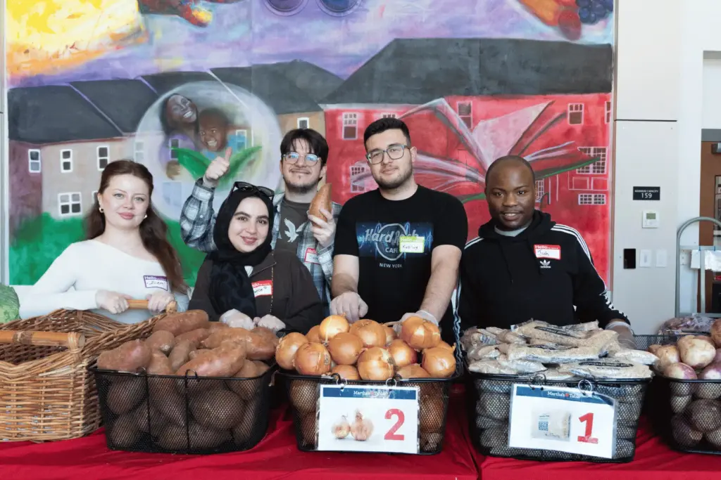 BAU Program Images (68) Volunteers smiling at a food drive table with baskets of potatoes and onions, colorful mural backdrop.