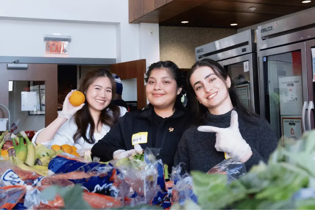 BAU Program Images (72) Three smiling people volunteering at a food bank, surrounded by fresh fruits and vegetables.