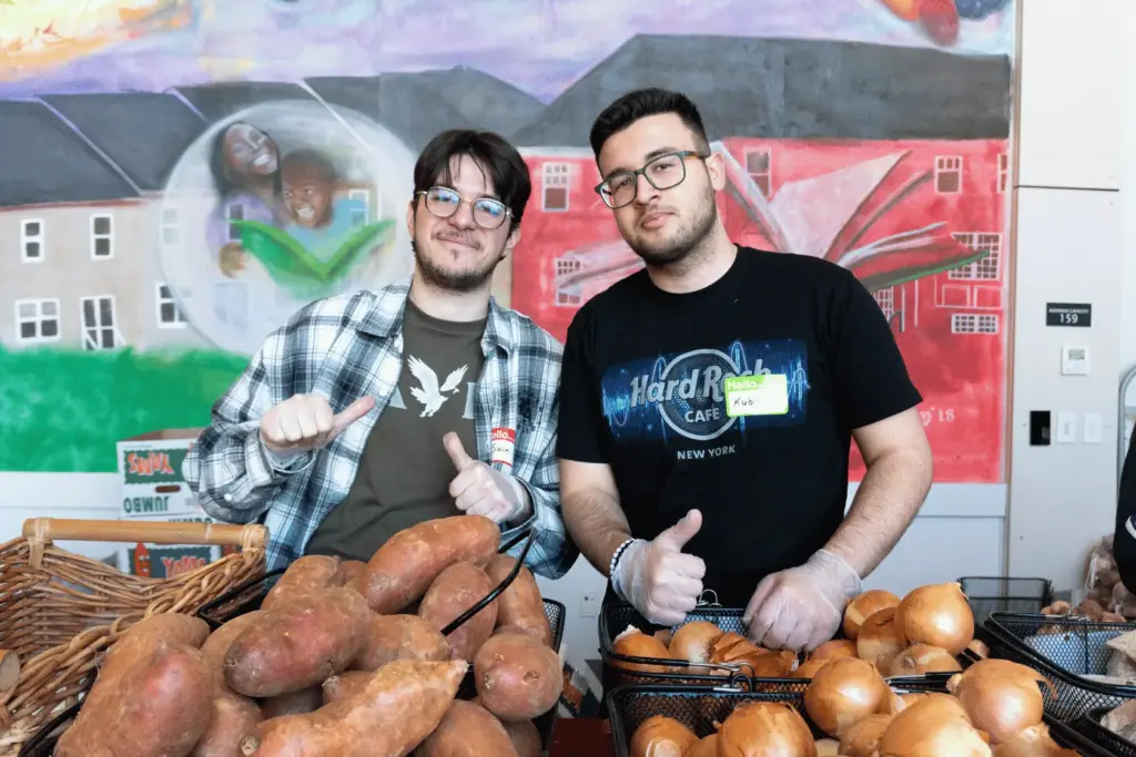 BAU Program Images (73) Two men giving thumbs up behind a display of sweet potatoes and onions at a market.