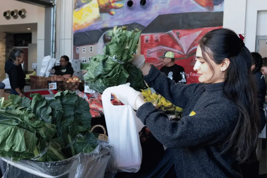 BAU Program Images (74) Woman packing fresh vegetables at a market with colorful mural in background.