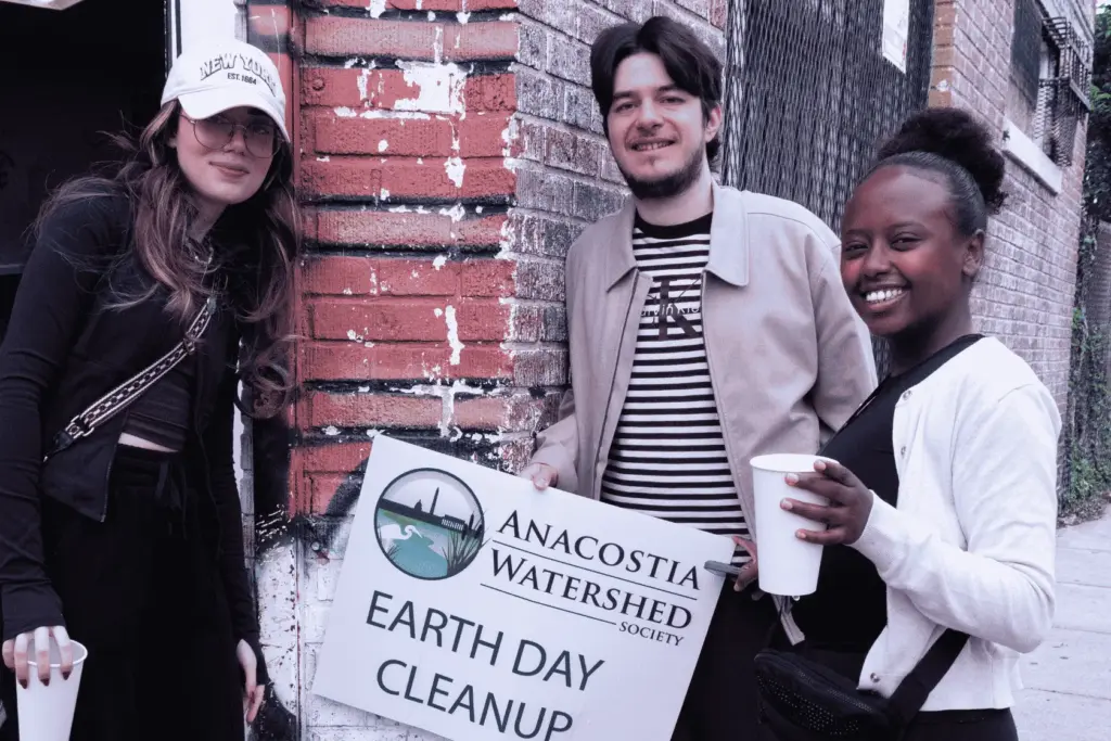 BAU Program Images (77) People at Anacostia Watershed Earth Day Cleanup event, holding a sign and cups, next to a brick wall.