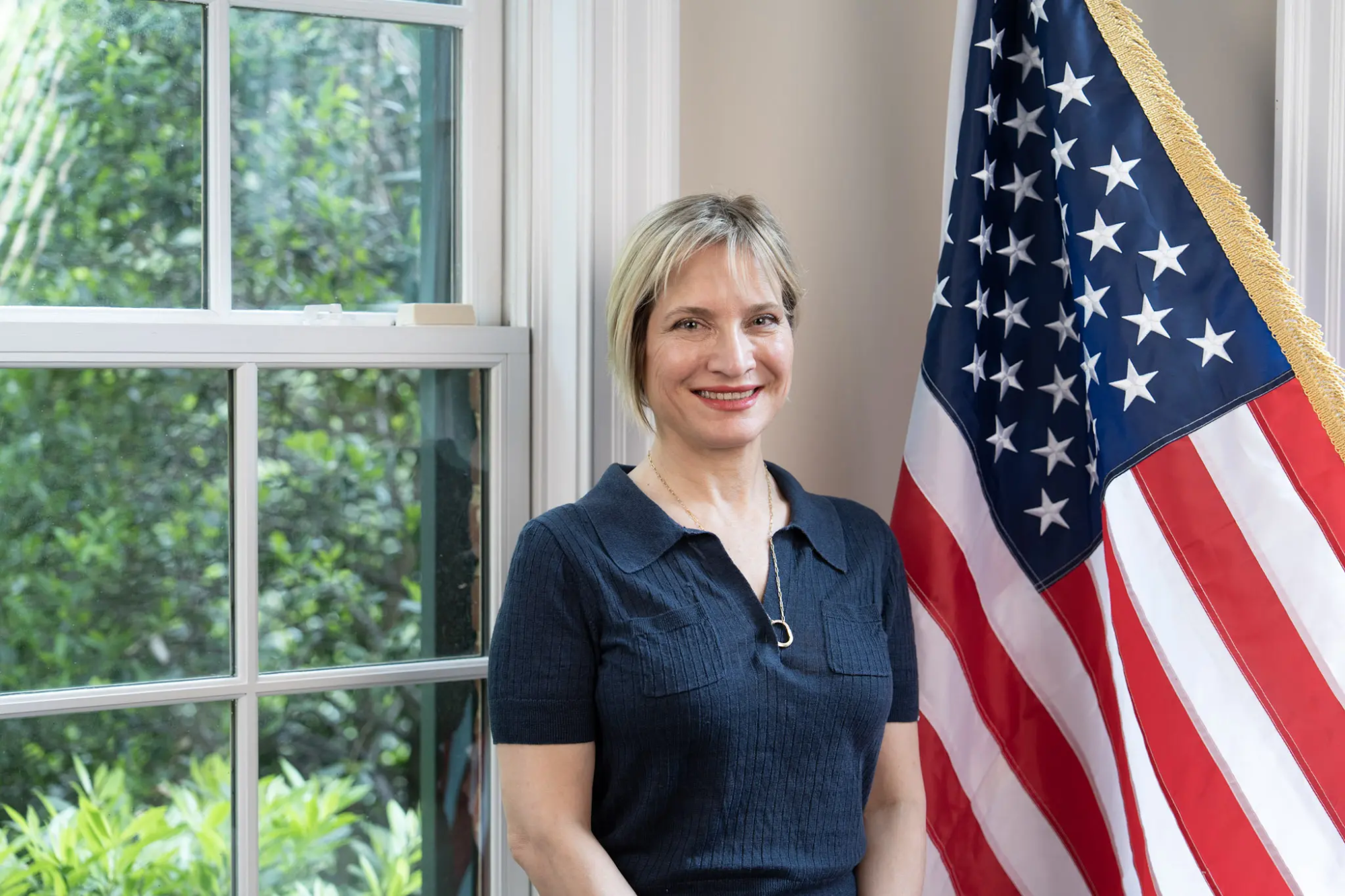Smiling woman standing next to an American flag by a window, showcasing patriotism and natural light.