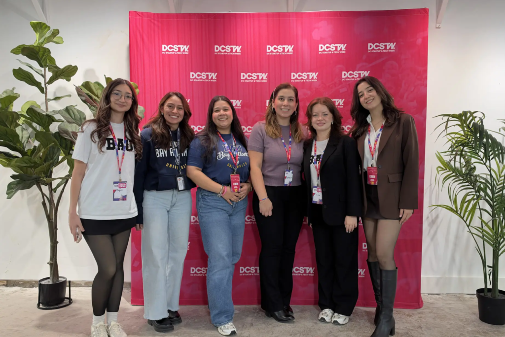 Group of six women smiling at a DCSTW event with a red backdrop and indoor plants.