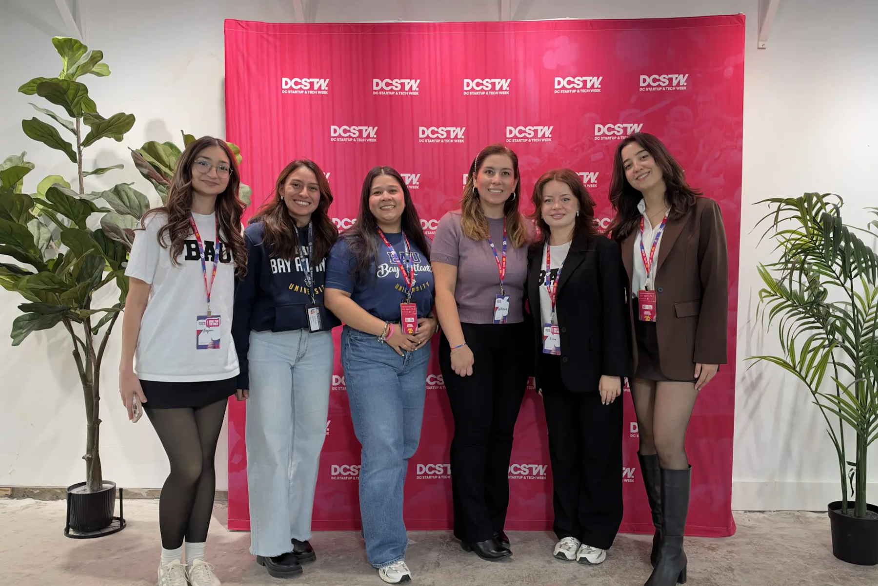 Group of six women smiling at a DCSTW event with a red backdrop and indoor plants.