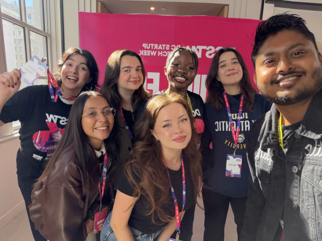 Group of diverse individuals smiling at a tech event with pink banner in the background.