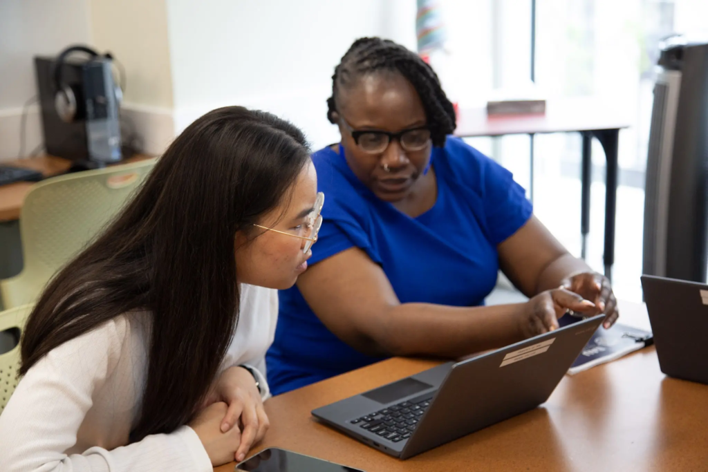 Two people collaborating on laptops in a classroom setting, focusing on a project together.
